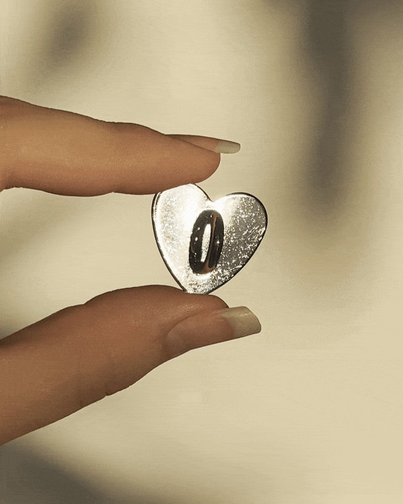 LoveBeats Heart Phone Charm Holder, a close-up of the polished silver heart metal anchor point for phone cases, held between two fingers.
