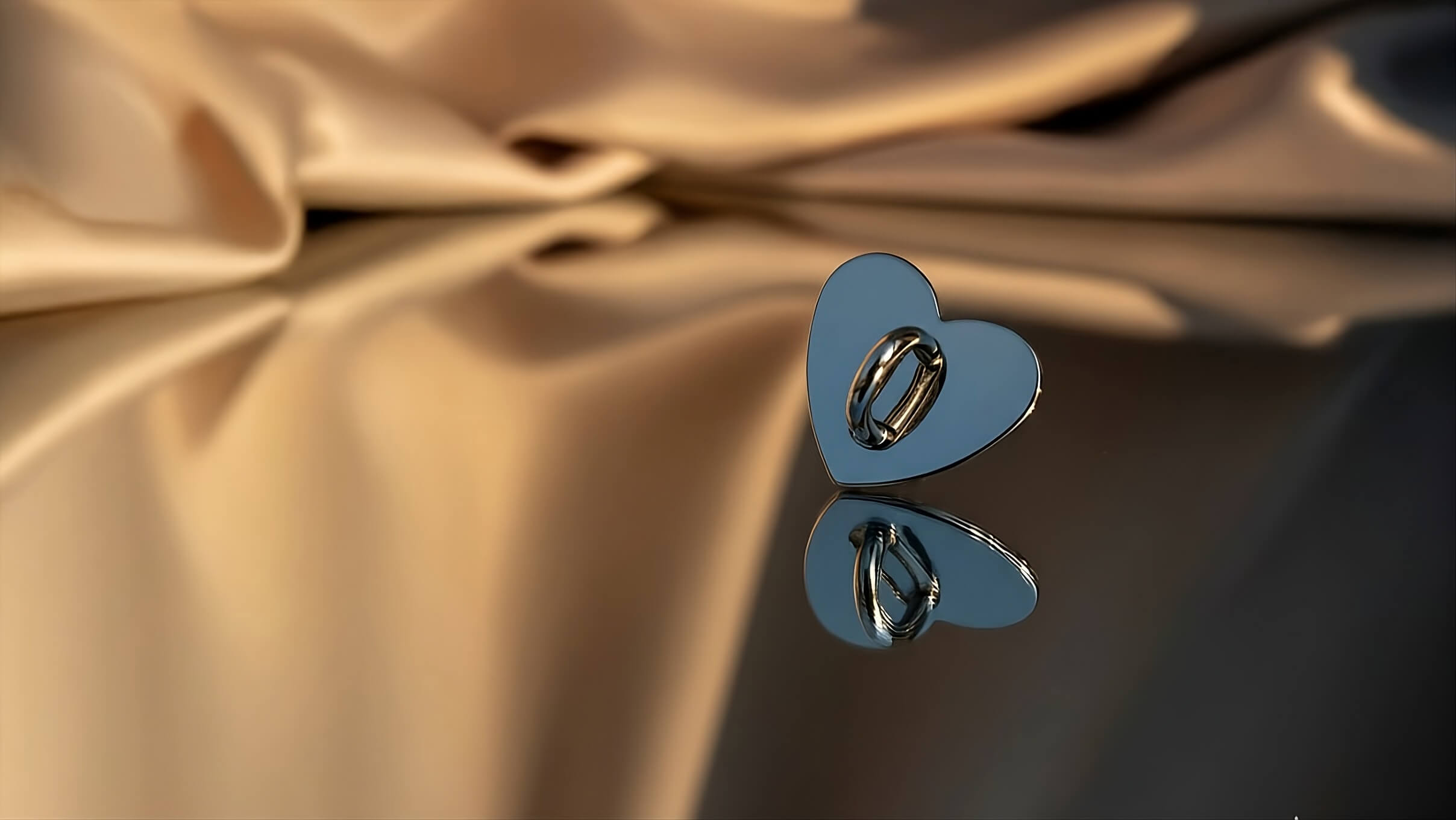 Close-up of a sophisticated silver heart-shaped phone charm holder. The polished metal anchor is designed to attach to a phone case for securing bag charms or lanyards. Luxury phone accessory by Love Beats